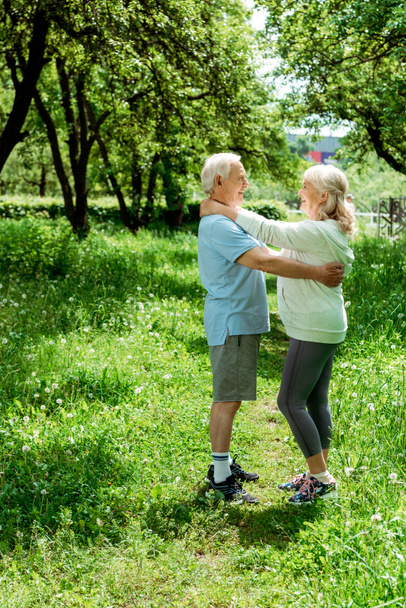улыбающийся пенсионер обнимает счастливую старшую жену, стоя в зеленом парке
  - Фото, изображение
