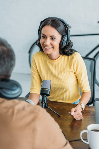 selective focus of smiling radio host in headphones looking at colleague while sitting near microphone in broadcasting studio - Фото, изображение
