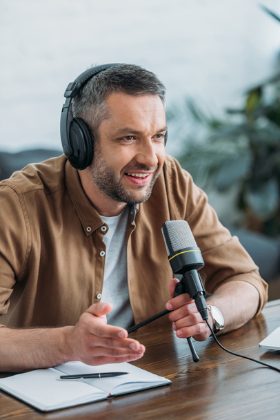 handsome radio host in headphones gesturing while speaking in microphone in radio studio - Fotoğraf, Görsel