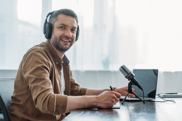 χαρούμενο ραδιοφωνικό ξενιστή στα ακουστικά που χαμογελούν στην κάμερα ενώ κάθονται κοντά στο μικρόφωνο και το φορητό υπολογιστή - Φωτογραφία, εικόνα