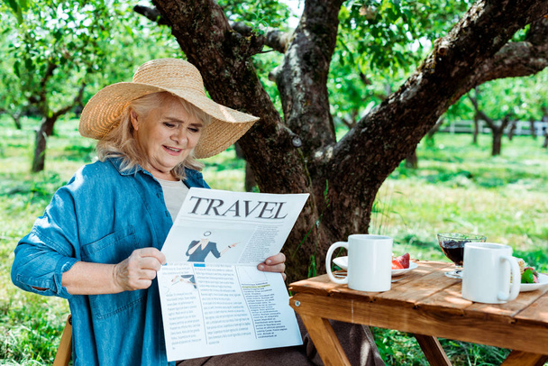 mulher sênior em chapéu de palha sentado perto da árvore e ler jornal de viagem
 - Foto, Imagem