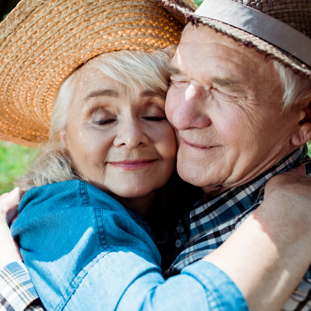 close up of cheerful senior woman hugging happy retired husband in straw hat  - Zdjęcie, obraz
