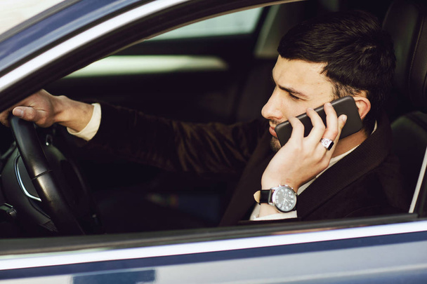 Ο νεαρός κουστουμάτος μιλάει τηλεφωνικώς στο αμάξι του. Bussines ματιά. Δοκιμή κίνησης του νέου αυτοκινήτου - Φωτογραφία, εικόνα