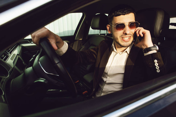 Young bussinesman in suit and black glasses speaks by phone in his car. Bussines look. Test drive of the new car - Foto, afbeelding