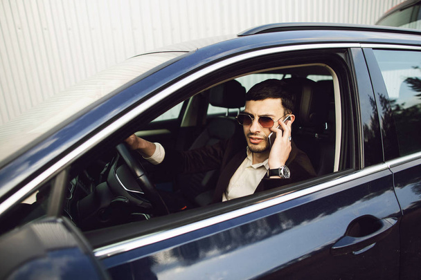 Joven hombre de negocios en traje y gafas negras habla por teléfono en su coche. Los negocios miran. Prueba de conducción del coche nuevo
 - Foto, imagen
