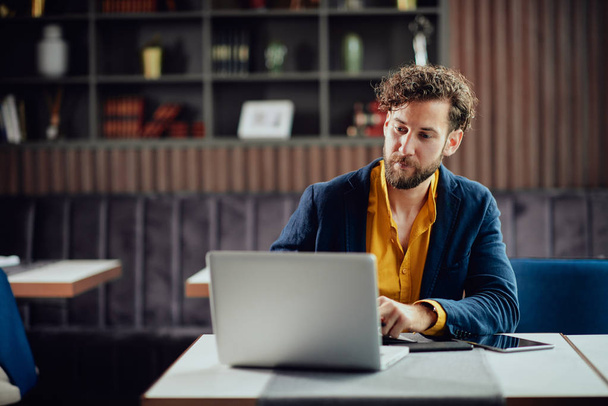Νεαρός σοβαρός γενειοφόρος επιχειρηματίας με έξυπνα casual ρούχα κάθεται στο καφέ και πληκτρολογώντας στο laptop. - Φωτογραφία, εικόνα