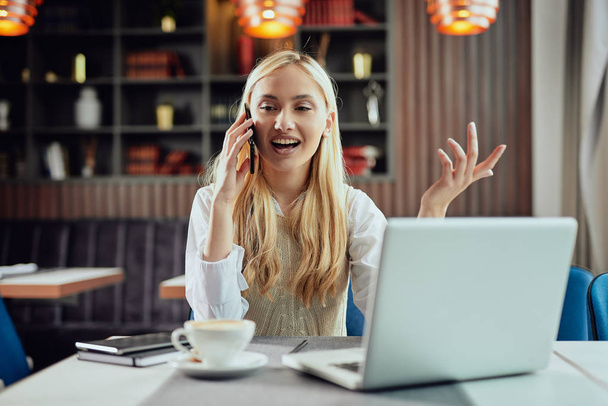Jovem mulher de negócios loira caucasiana alegre vestida inteligente casual sentado no café e usando telefone inteligente
. - Foto, Imagem