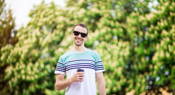 Un joven atractivo con cabello oscuro y gafas de sol está caminando por la calle con una taza de café en un día soleado. barbudo hipster chico con ropa casual disfruta relajarse en un fin de semana de verano
. - Foto, imagen