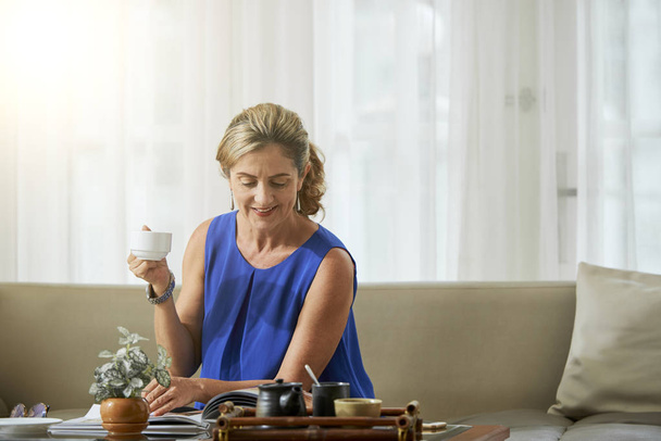 Mulher madura relaxando no sofá com xícara de café e lendo uma revista na sala de estar em casa
 - Foto, Imagem