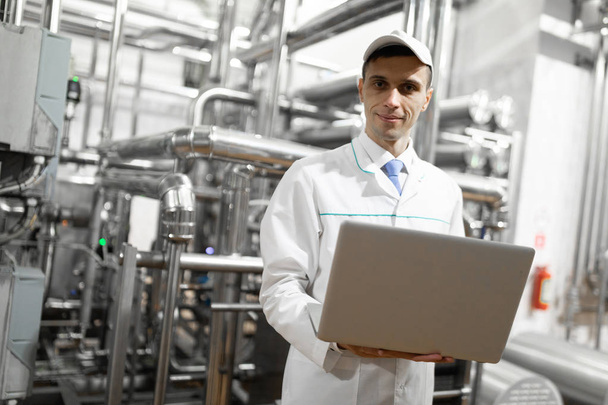 Portret człowieka w białym szacie i czapkę stojącą w dziale produkcyjnym fabryki mleczarskiej z laptopem - Zdjęcie, obraz