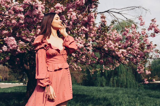 a young brunette maid in the park, with sunglasses, mingling with a blooming tree, dressed in a pink and sexy summer dress - Fotoğraf, Görsel