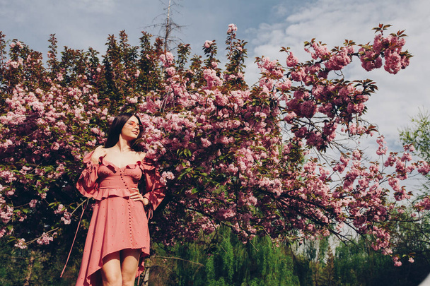 a young brunette maid in the park, with sunglasses, mingling with a blooming tree, dressed in a pink and sexy summer dress - Photo, image