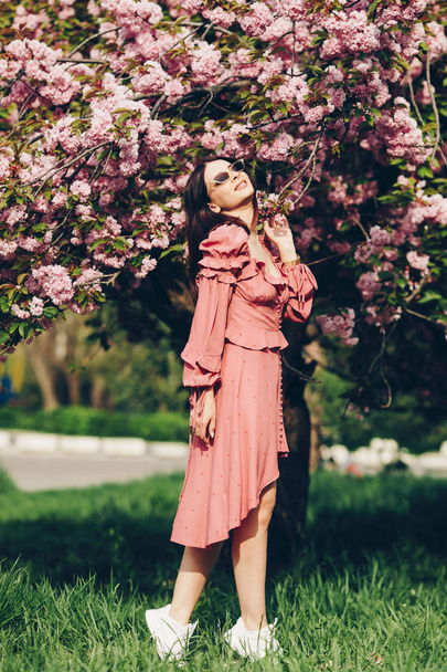 Mladá brunetka v parku, s slunečními brýlemi, s rozkvetlou větví, oblečenou v růžových a sexy letních šatech - Fotografie, Obrázek
