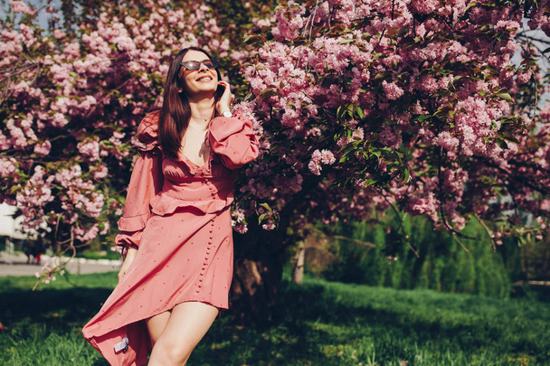 a young brunette maid in the park, with sunglasses, mingling with a blooming tree, dressed in a pink and sexy summer dress - Photo, image