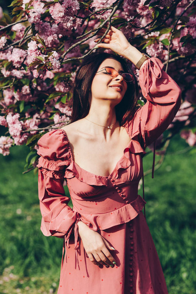 a young brunette maid in the park, with sunglasses, mingling with a blooming tree, dressed in a pink and sexy summer dress - Photo, Image