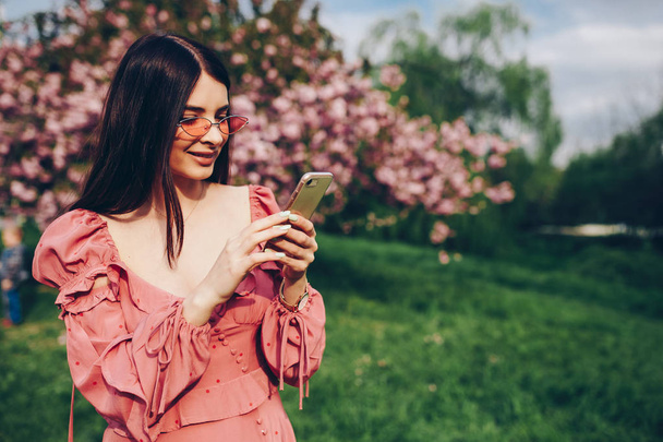 молода леді в парку в короткому рожево-червоному рушничку, який тримає телефон і розмовляє, пише текстове повідомлення, розмовляє онлайн
 - Фото, зображення