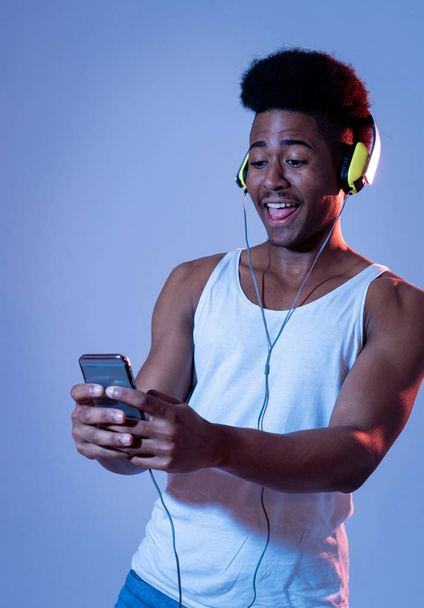 Strong african american young man listening to music in smartphone app looking for best song online with headphones singing and dancing. Portrait with disco dance floor light in Music and technology. - Foto, Imagen