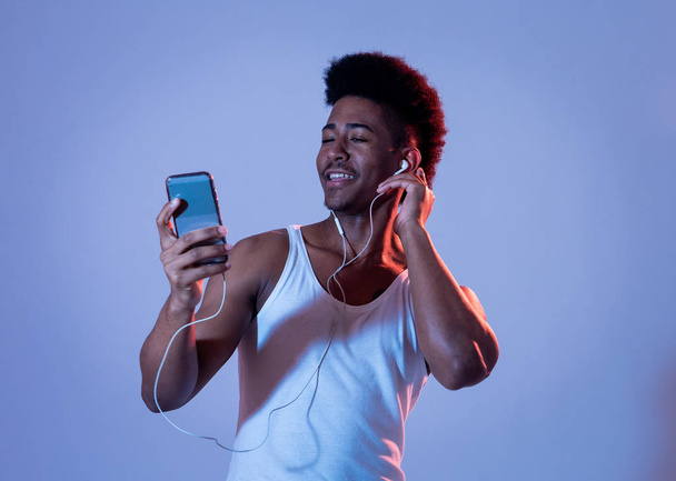 Strong african american young man listening to music in smartphone app looking for best song online with headphones singing and dancing. Portrait with disco dance floor light in Music and technology. - Foto, Imagen