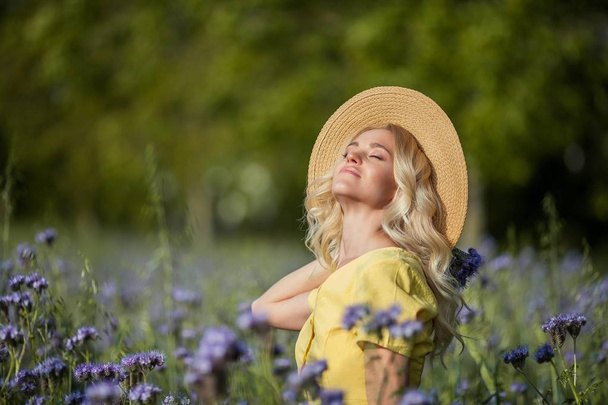 Young beautiful woman blonde in a hat walks through a field of purple flowers. Summer. - Foto, afbeelding