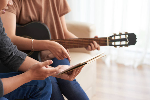 Boční pohled na dvojici sedícího pohromadě na pohovce, muž držící tablet a ženu s akustickou kytarou ukazující na obrazovce na rozmazané pozadí  - Fotografie, Obrázek