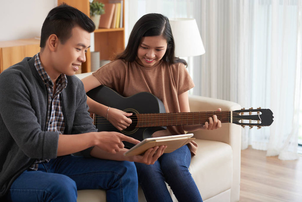 Vista lateral del alegre hombre asiático en ropa casual mostrando a la joven atractiva morena sentada con la tableta de guitarra y apuntando a la pantalla, ambos sonriendo sentados en el sofá
 - Foto, imagen