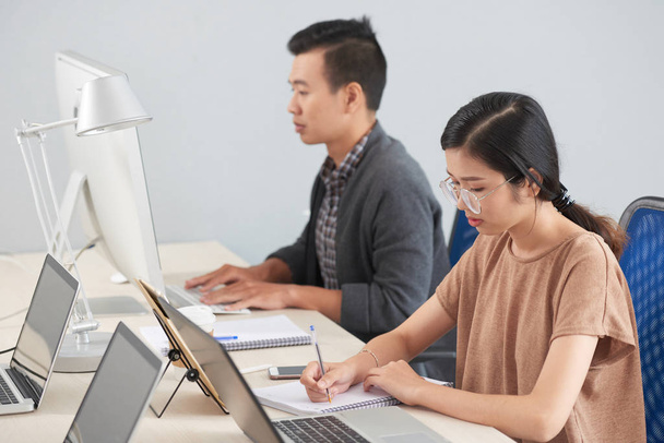 ノートパソコンとぼやけた背景にコンピュータで作業している男性と机に座ってメモ帳に書くことに集中メガネで魅力的なアジアのブルネットの女性の側面図  - 写真・画像