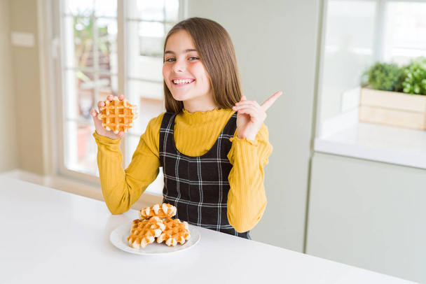 Beautiful young girl kid eating Belgian waffle pastry very happy pointing with hand and finger to the side - Foto, imagen