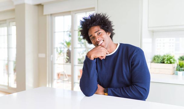 junger afrikanisch-amerikanischer Mann in lässigem Pullover, der zu Hause sitzt und selbstbewusst in die Kamera lächelt, mit verschränkten Armen und erhobener Hand am Kinn. Positives Denken. - Foto, Bild