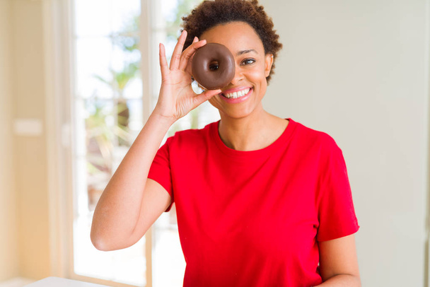 Młoda Afroamerykanka jedząca pączka czekoladowego ze szczęśliwą twarzą na stojąco i uśmiechnięta z pewnym siebie uśmiechem pokazującym zęby - Zdjęcie, obraz