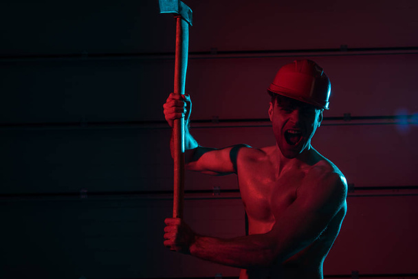 screaming sexy shirtless fireman in helmet holding flat head axe in darkness - Foto, imagen