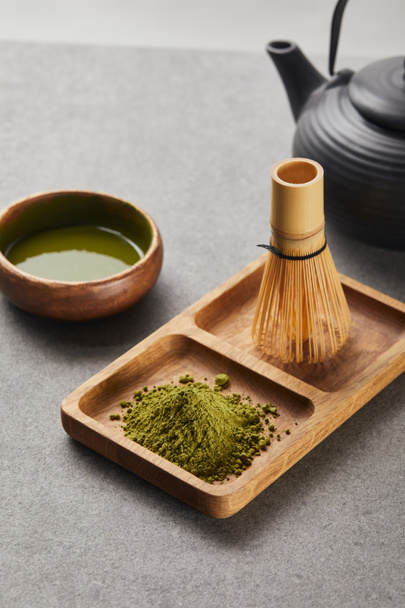 selective focus of green matcha powder and bamboo whisk on wooden board near black teapot and bowl with tea - Foto, afbeelding