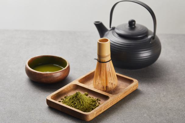 selective focus of green matcha powder and bamboo whisk on wooden board near black teapot and bowl with tea - Foto, afbeelding