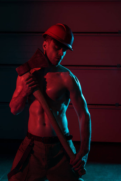 sexy shirtless fireman in protective helmet holding flat head axe in darkness - Фото, изображение