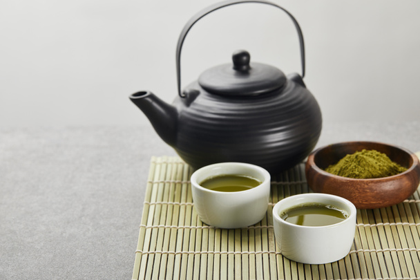 selective focus of green matcha powder in wooden bowl near black teapot and cups with tea on bamboo table mat - Фото, зображення