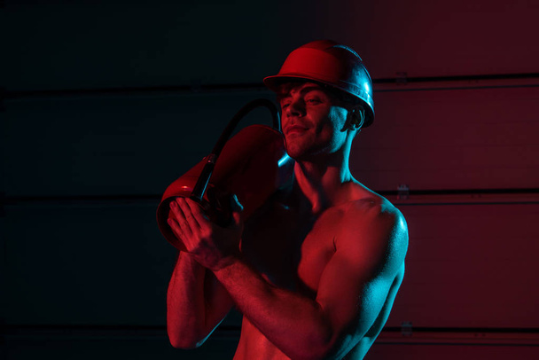 sexy shirtless fireman in protective helmet holding fire extinguisher in darkness - Photo, image