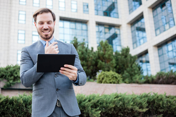 glücklicher junger Geschäftsmann, der vor einem Bürogebäude an einem Tablet arbeitet - Foto, Bild