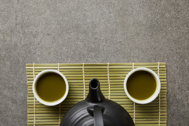 top view of white cups with matcha and black teapot on green bamboo table mat - Photo, Image