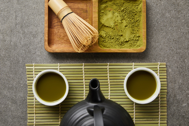top view of white cups with tea and black teapot on green table mat near bamboo whisk and matcha powder on board - 写真・画像