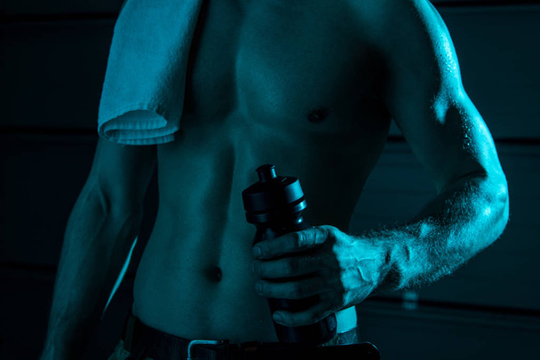 partial view of sexy shirtless man with towel holding sport bottle in darkness - Foto, imagen