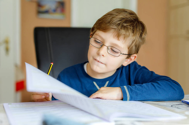 Portret van schattige school jongen jongen met bril thuis maken van huiswerk. Weinig geconcentreerd kind schrijven met kleurrijke potloden, binnenshuis. Basisschool en onderwijs - Foto, afbeelding