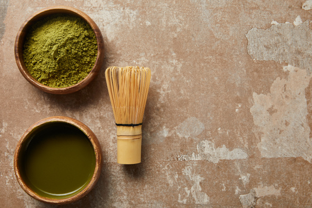 top view of matcha tea and green powder in wooden bowl with bamboo whisk on aged surface - Foto, Bild
