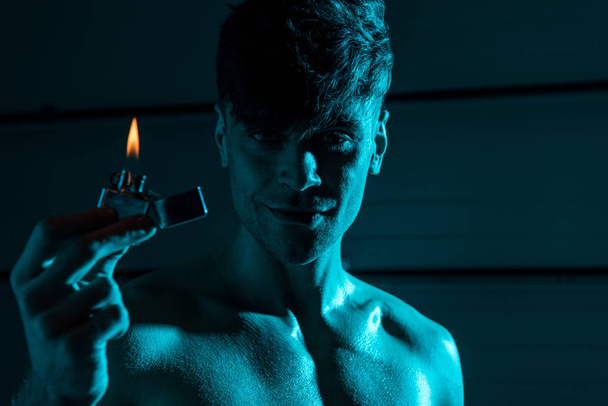 sexy shirtless muscular man holding lighter and smiling in darkness - 写真・画像