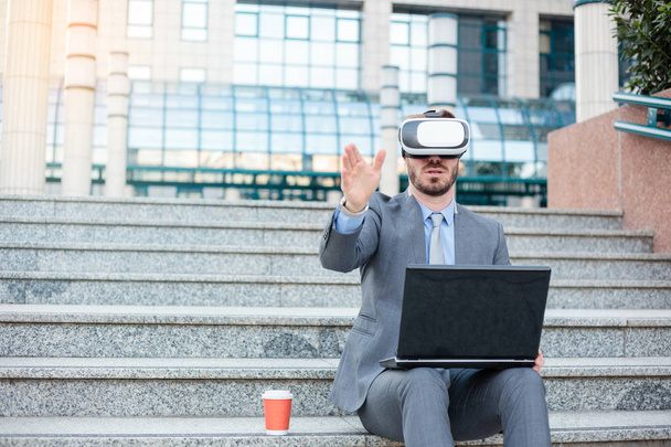 Успішний молодий бізнесмен використовує VR окуляри і робить жести рук, працюючи на ноутбуці перед офісною будівлею
 - Фото, зображення