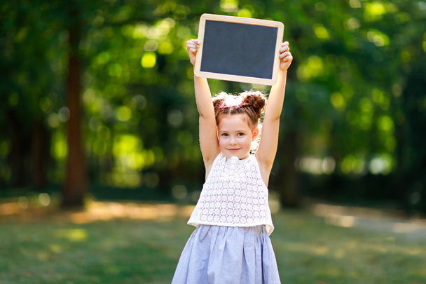 Glückliches kleines Mädchen mit leerem Kreidetisch in der Hand. Schulkind am ersten Tag der Grundschule. Gesundes entzückendes Kind im Freien, im grünen Park. Kopierraum auf dem Schreibtisch - Foto, Bild