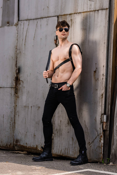 Ganzkörperansicht von sexy Mann ohne Hemd in schwarzer Sonnenbrille und Schwertgürtel mit Hand in Tasche - Foto, Bild
