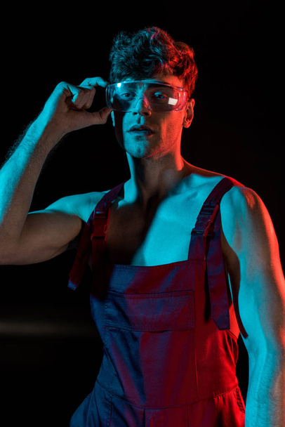 sexy shirtless fireman in overall touching protective goggles on black - Fotoğraf, Görsel