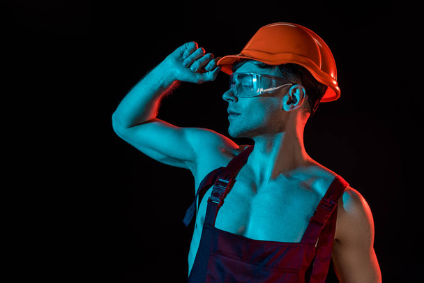 sexy fireman in overall, hardhat and protective goggles isolated on black - Zdjęcie, obraz