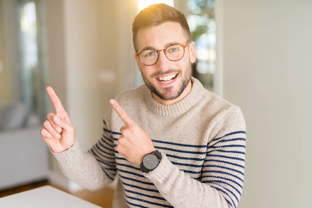 Νέος όμορφος άντρας φορώντας γυαλιά στο σπίτι χαμογελώντας και κοιτώντας την κάμερα που σημαδεύει με δύο χέρια και δάχτυλα στο πλάι. - Φωτογραφία, εικόνα