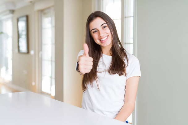 schöne junge Frau in legerem weißen T-Shirt mit glücklichen Daumen nach oben Geste mit der Hand. zustimmender Ausdruck, der mit Erfolg in die Kamera blickt. - Foto, Bild