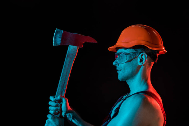 side view of sexy fireman in hardhat and protective goggles holding flat head axe isolated on black - Foto, imagen